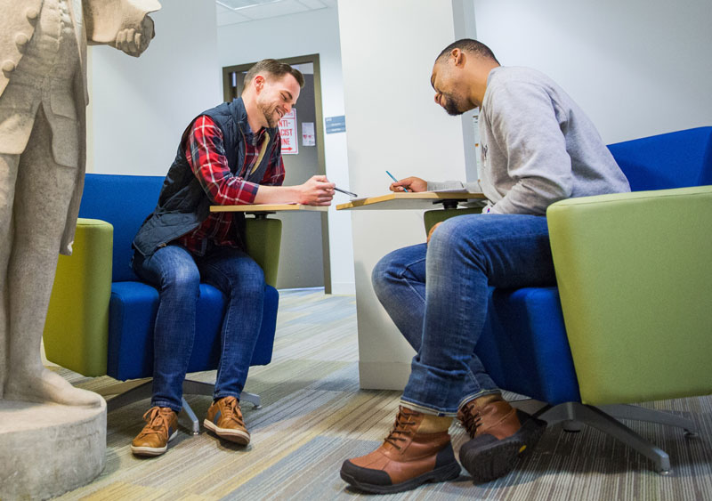 Two men wearing casual shirts and blue jeans are sitting across from each other in a student lounge area, smiling and looking at material on the desks in front of them