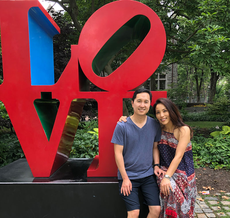 A couple wearing casual outfits in red and blue pose while leaning on the LOVE statue on Penn’s campus. Fresh foliage is seen all around in the background.