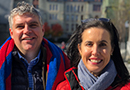 A smiling man and a woman, both in red winter coats, stand outside.