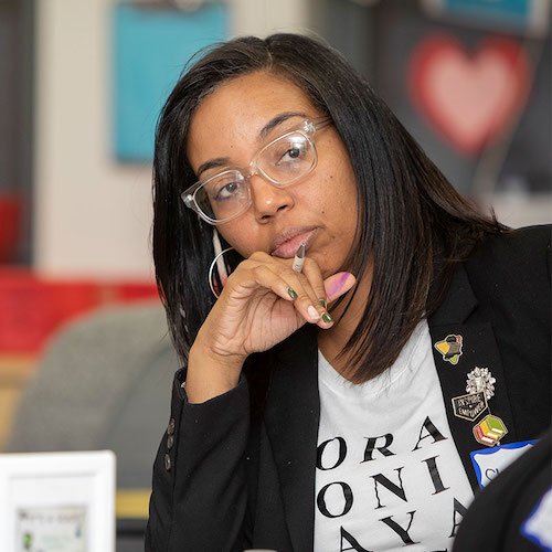 A teacher sitting at a table tilts her head to one side in thought. Around her is a colorful classroom.
