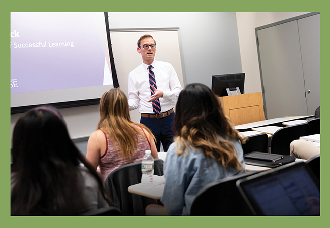 A man wearing glasses and professional attire stands and teaches in front of a classroom.