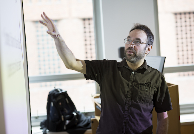 Ryan Baker at the front of a classroom gesturing toward information projected onto a screen. 