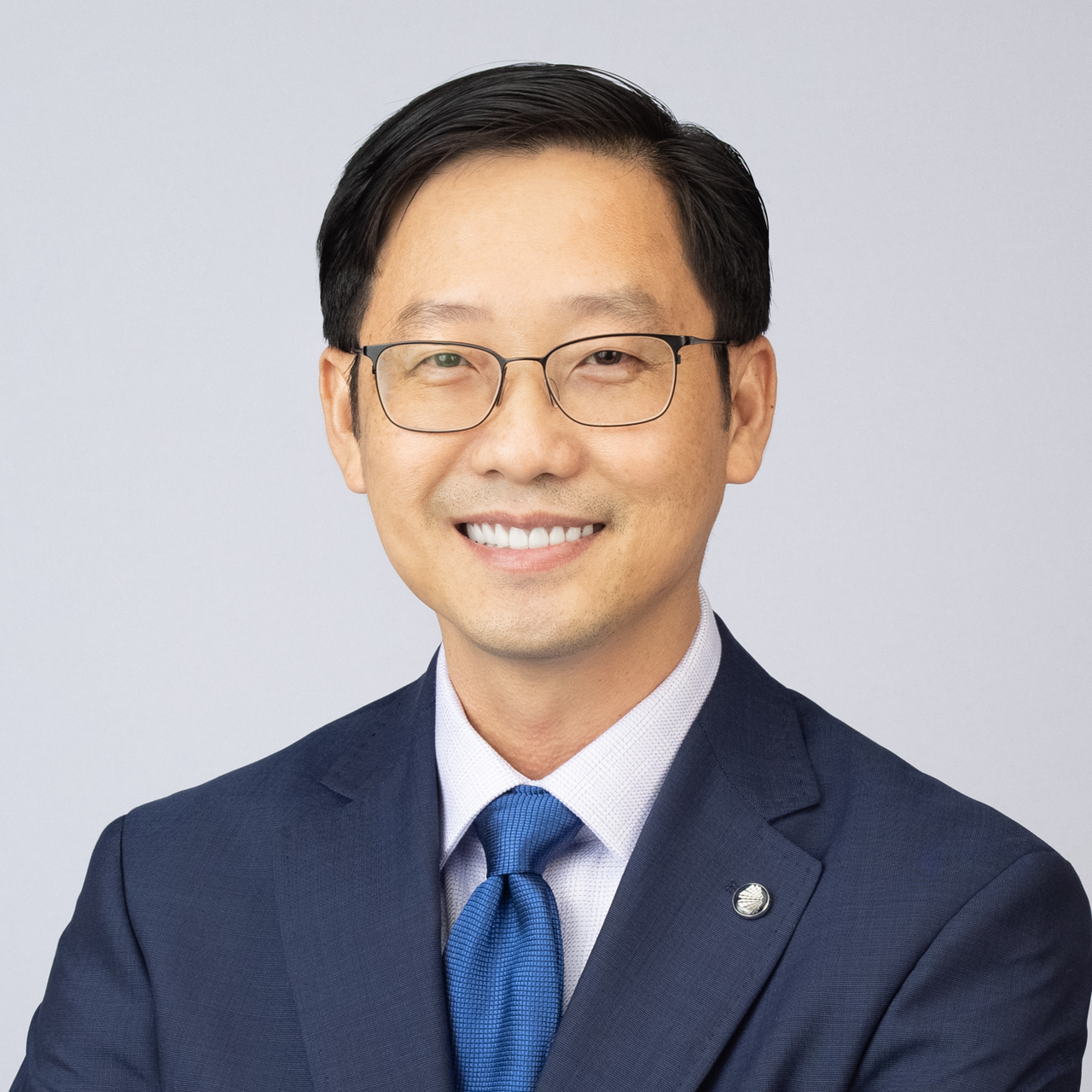 An Asian man wearing a blue suit and blue tie with glasses smiles into the camera