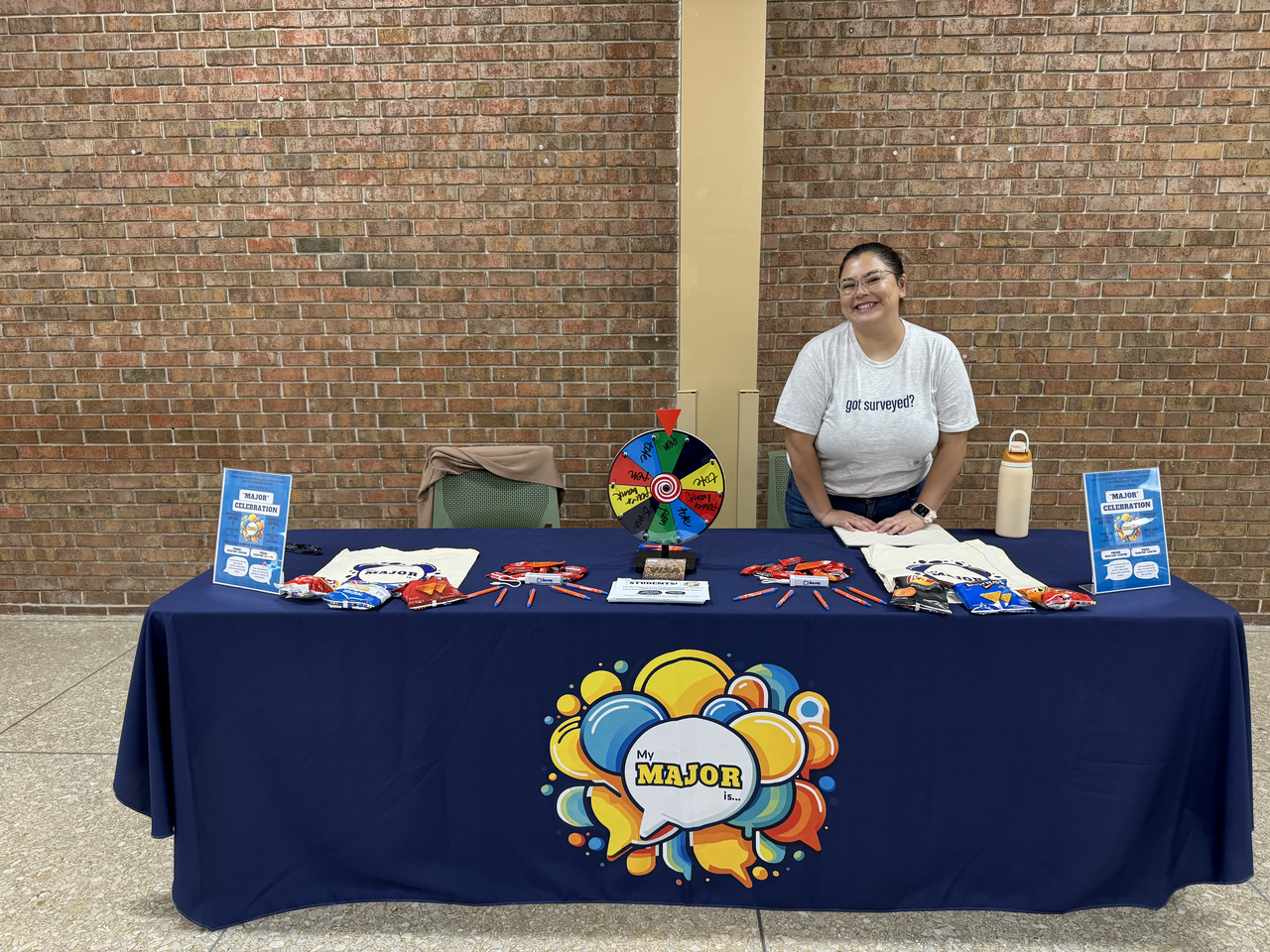 Estefanie stands behind a table covered in pens and other swag, wearing a shirt that says "got surveyed?"
