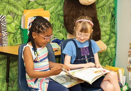 wo school girls sitting next to each other on blue chairs are smiling and flipping through pages of a large book with colorful illustrations.