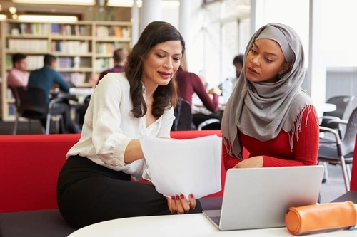Adult female pointing to a paper she is holding while sitting with a younger female student who is wearing a scarf on her head.