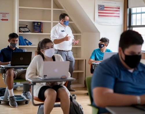 Four masked students sitting at their desks with laptops open as a teacher talks to one of them.