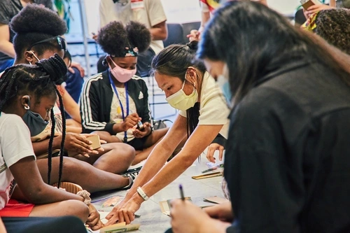A masked teacher is explaining something to students sitting on the floor.