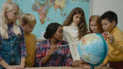 Teacher pointing at a globe on her desk with children surrounding her.