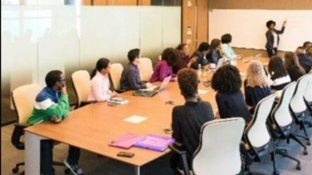 Classroom with students sitting around a large boardroom style table looking at the a person presenting in front of a whiteboard.