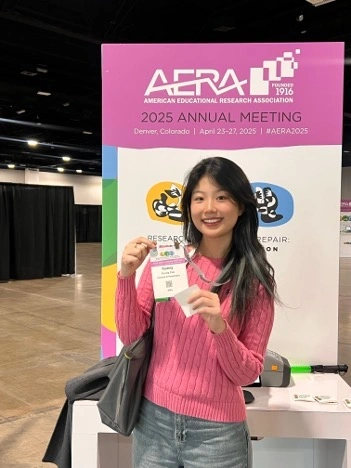 a young asian woman holds a name tag smiling while standing in front of a banner stating "AERA 2025 Annual Meeting"