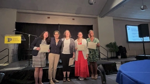 Five women pose with award certificates in front of a stage and a podium that has a yellow sign on it that reads “Community College of Philadelphia”.