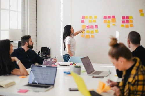 Female pointing at colorful post-its on a whiteboard, in front of colleagues sitting around a conference table.