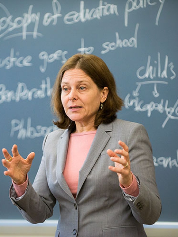 A professor addresses a class, standing in front of a blackboard with white chalk writings related to philosophy and education.