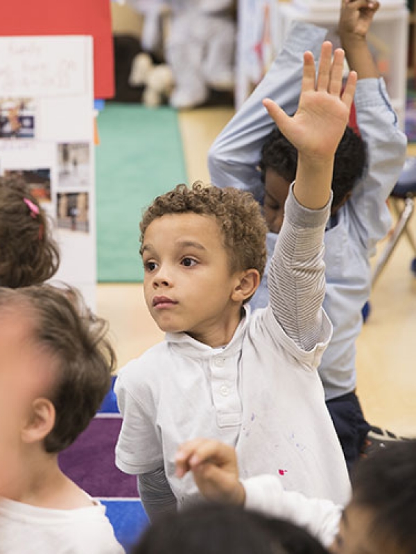 Elementary school children sit on the floor with their hands raised inside a colorful classroom.