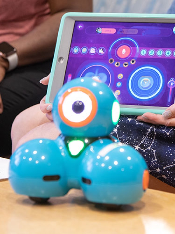 A blue robotic device with three spheres sits on a table. Behind it, an individual holds up a tablet device showing illuminated circles.