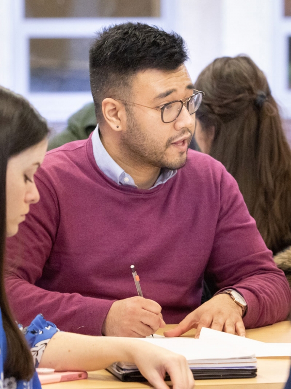 Two graduate students are sitting at a desk in a classroom, with other students seen in the background. One student takes notes while conversing, the other student looks down.