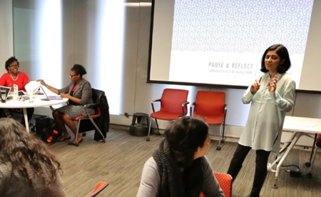 Students sit around four round tables as Ameena Ghaffar - Kucher leads the class with a screen behind her with a projection on it that reads “Pause and Reflect.”