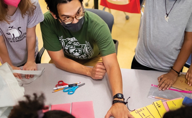 A seated teacher, wearing a face mask and glasses, is surrounded by several masked elementary students and reaches across a table to engage with a student's paper.