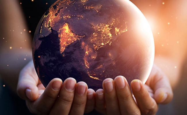 A person holds the planet earth in their cupped hands. The earth is illuminated by city lights and the sun rising on the right side of the planet. 