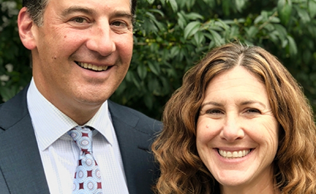 A man in a suit and tie and a woman in a black blouse smiling while standing outdoors