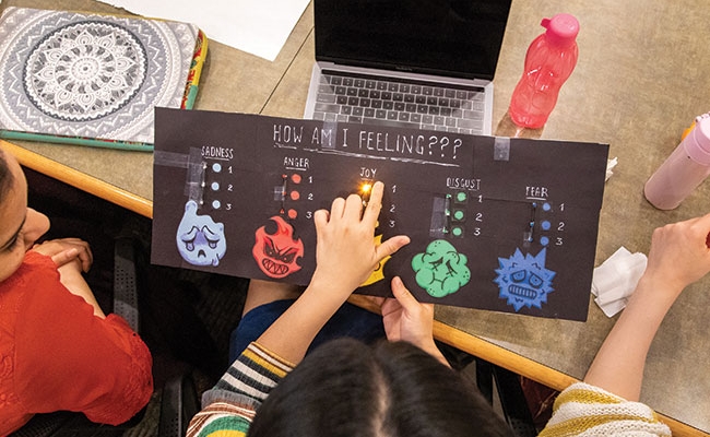 Aerial view of a student holding a colorful chart with a list of emotions and a light shining in the middle underneath the word “Joy.”