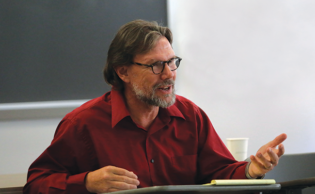 Photo of Michael Nakkula in a red button-up shirt and glasses sitting at a desk and teaching
