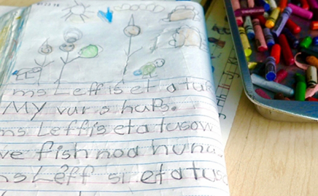 A folded composition book with children’s writing and drawings lies next to a tray of crayons 