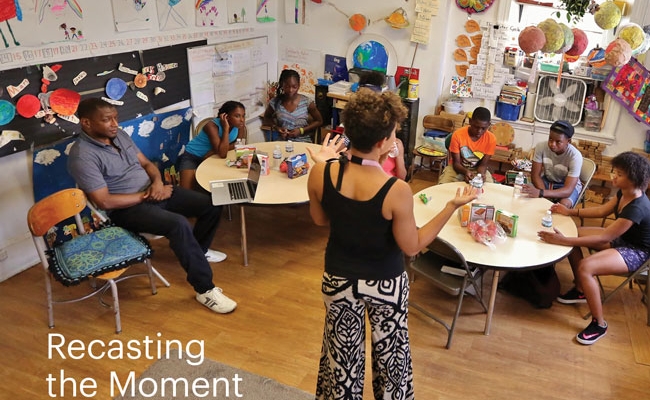 Elementary school students sit at round tables in a classroom. Behind them are colorful decorations, classroom projects, and school supplies. A woman stands at the front of the room and a man sits to the left. A headline on the image reads, “Recasting the Moment.”