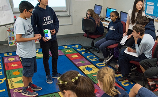 Students sitting in computer room, facing two students presenting an app they created on a tablet. 