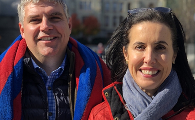 A smiling man and a woman, both in red winter coats, stand outside.