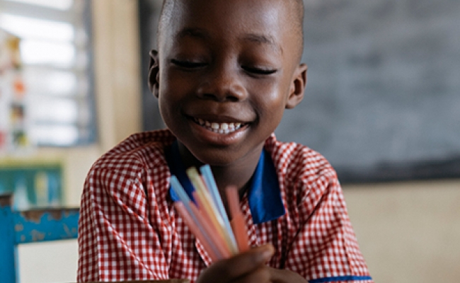 A child in a classroom in Ghana smiles while holding a handful of pens