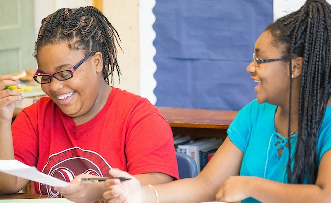 Two students sitting in a classroom laughing while collaborating on a paper