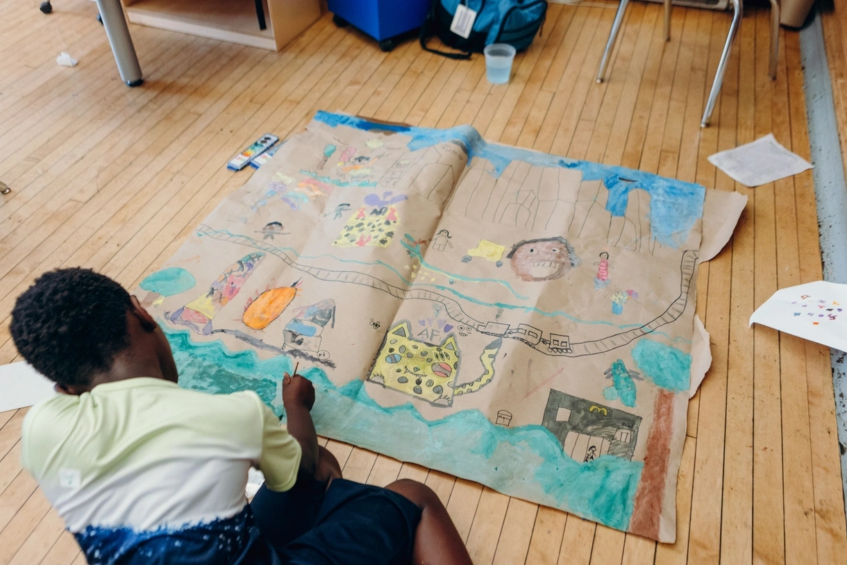A Henry C. Lea Elementary School student sits on the wood floor in the foreground, painting with teal watercolor on a large class art project on brown construction paper featuring people, vehicles, animals, a train, a McDonald's, a pumpkin, a rocketship, trees, skyscrapers, and more.