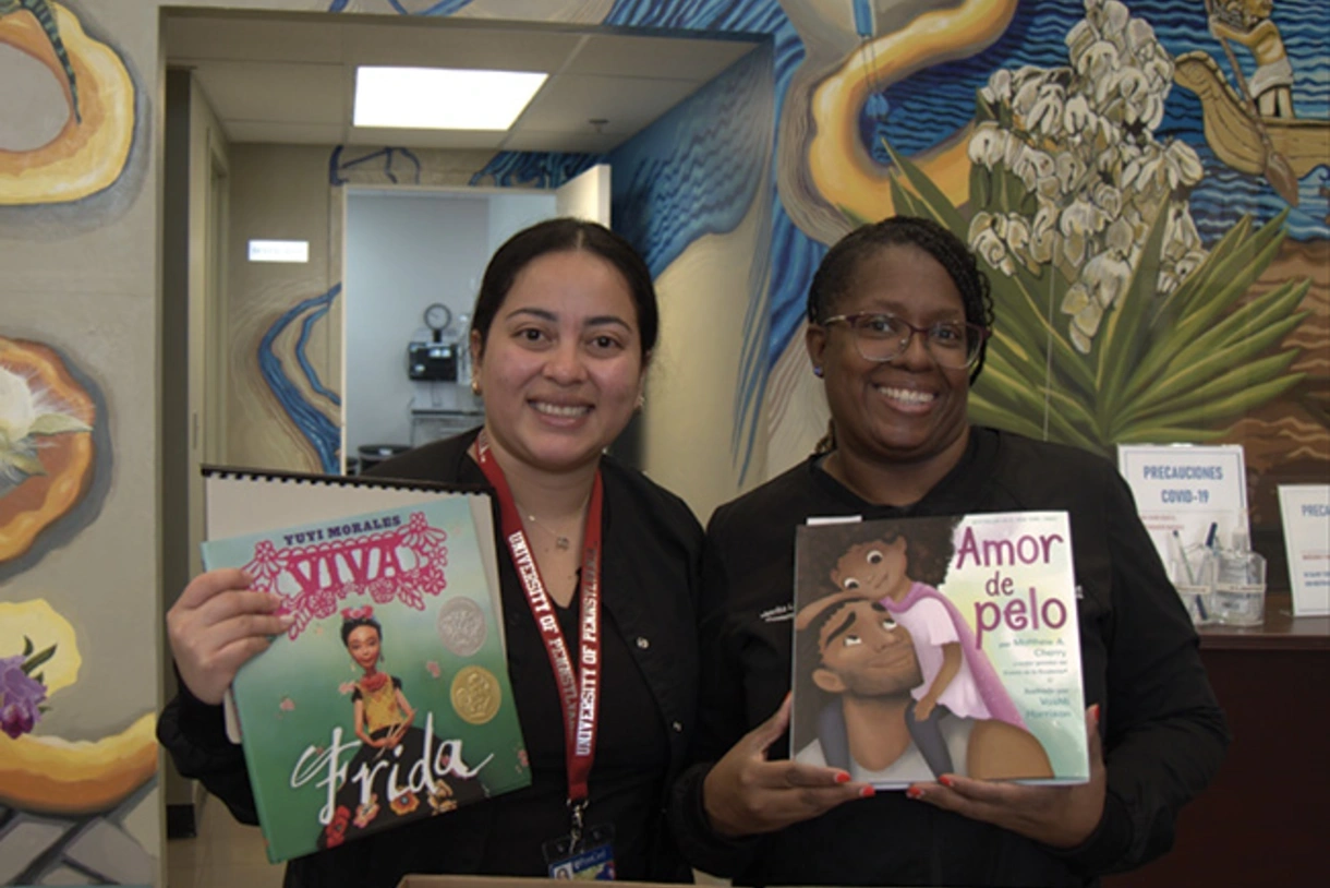 Two volunteers stand in the Puentes de Salud medical clinic with a mural painted on the wall behind them, holding children’s books in Spanish donated by A Book a Day