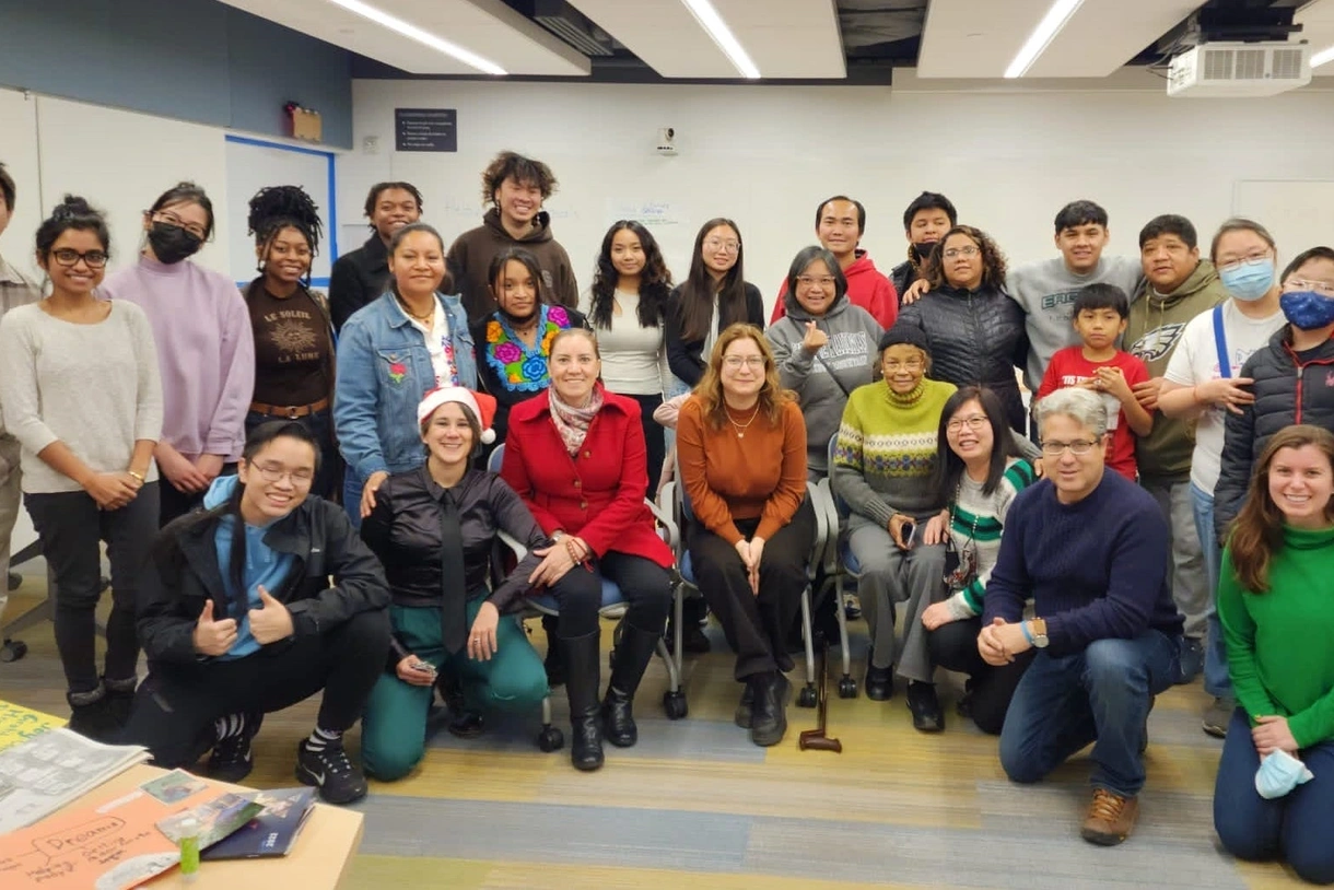 A large multicultural group of 27 youth, parents, and other adults pose together in a semicircle in a classroom, smiling.
