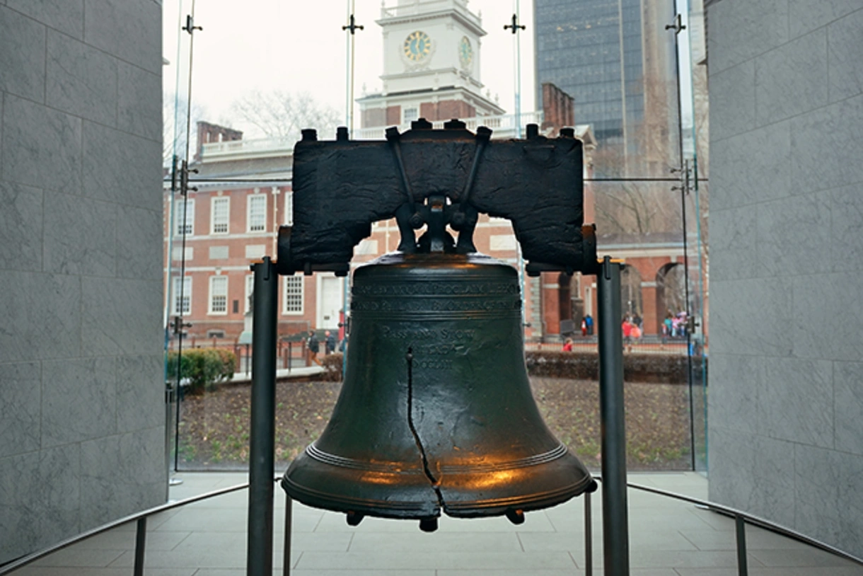 The Liberty Bell in Philadelphia appears between two gray walls, with traditional and modern buildings seen through a window behind it.