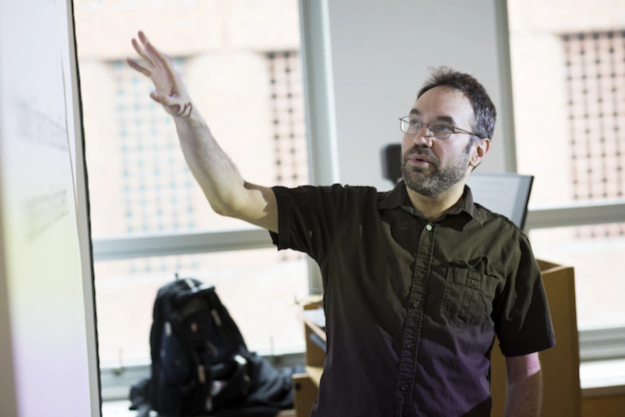 Ryan Baker at the front of a classroom gesturing toward information projected onto a screen.