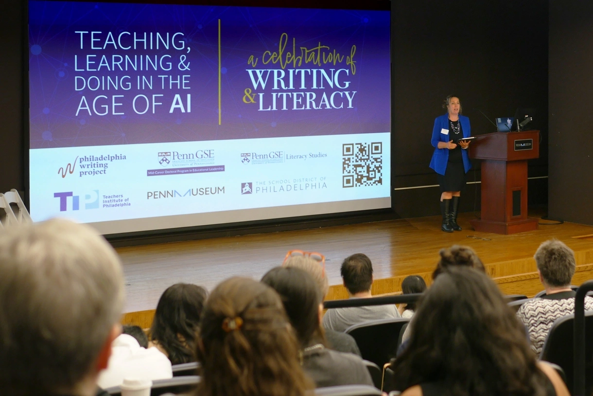 Amy Stornaiuolo speaks into a microphone on stage in front of an audience at the 2024 Celebration of Writing and Literacy with the conference logo on a screen behind her.