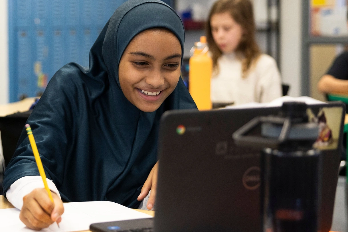 An elementary student in a hijab laughs while writing with a pencil and paper and looking at a laptop.
