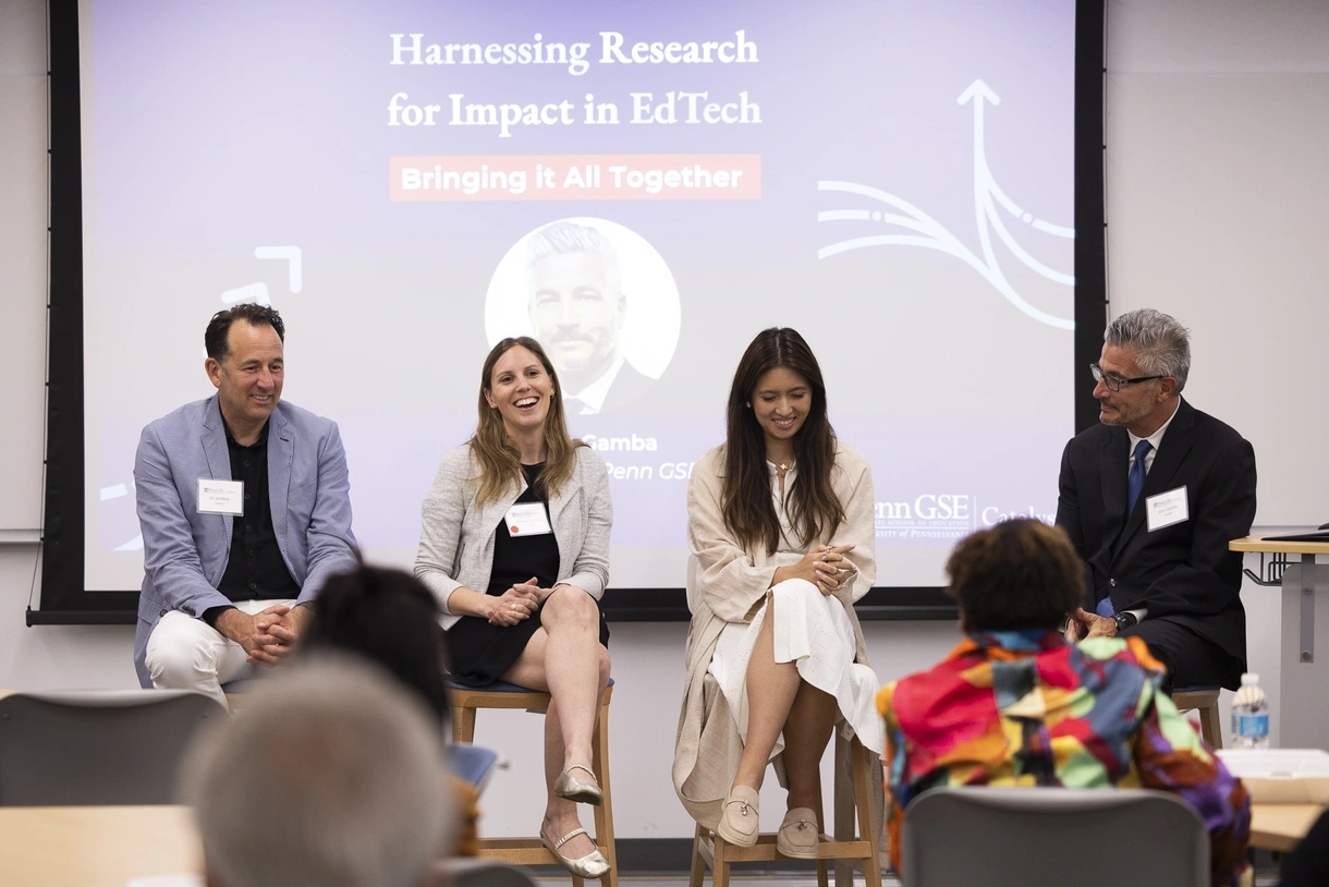 Four people sit on high chairs with their legs crossed and smiling in front of a room of people. A blue screen behind them reads "Harnessing Research for Impact in EdTech: Bringing it All Together"