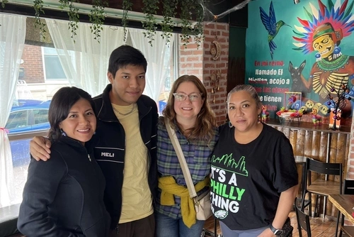 Four people stand together with their arms around one another in a restaurant, smiling at the camera. Behind them is a window on one wall and a mural of an Aztec god on another.