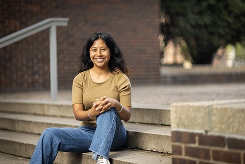 Penn GSE student Meresa García sits outside on cement steps in front of a brick wall on the University of Pennsylvania campus wearing a tan t-shirt and blue jeans, smiling at the camera