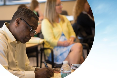 An adult student writes notes in a classroom. The photo is cropped with a semicircular edge against a blue-sky background.|