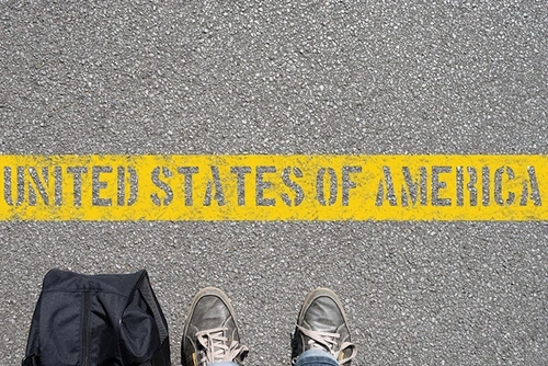 Two feet wearing shoes are pictured next to a black bag, at the bottom half of an image of a road with a thick yellow line on which “United States of America” is written.