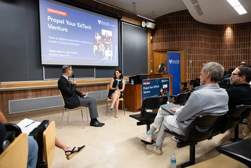 Penn GSE Dean Katharine Strunk, seated in a chair in front of an audience in a lecture hall, speaks into a microphone as Vice Dean of Innovative Programs and Partnerships Michael Golden listens, seated in a chair next to her. A screen behind them reads "Catalyst Summit: Propel Your EdTech Venture | How to deploy innovative solutions to tackle critical challenges."