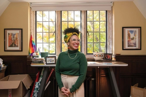 Krista Cortes stands, hands folded in front of her, laughing in the middle of her office surrounded by decorations showcasing Latin American culture.