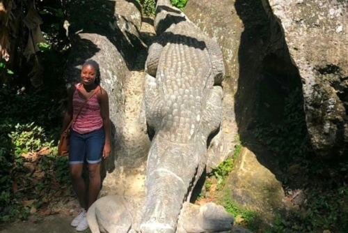 Angela Crumdy stands beside a large statue of an alligator biting another animal, surrounded by trees and rocks in Guantánamo, Cuba.