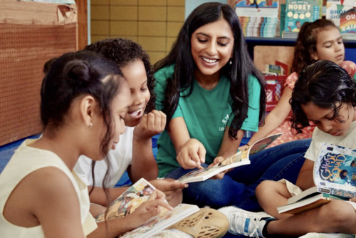 Rina wears a green t-shirt and sits on a rug with four children who are all reading and engaging together. 
