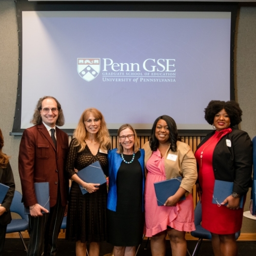 Dean Grossman stands in the middle of six smiling Education Alumni Award winners in front of a screen with the Penn GSE logo on it. 